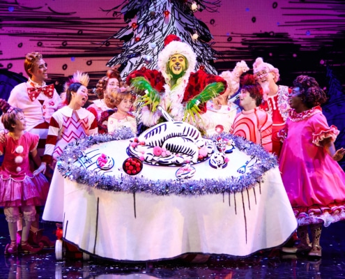A green furry creature dressed in a santa suit carves into an illustrated turkey on top of a large, round table. Other colorful actors in pink costumes surround the table