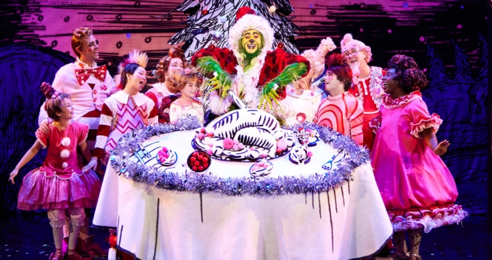 A green furry creature dressed in a santa suit carves into an illustrated turkey on top of a large, round table. Other colorful actors in pink costumes surround the table