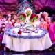 A green furry creature dressed in a santa suit carves into an illustrated turkey on top of a large, round table. Other colorful actors in pink costumes surround the table