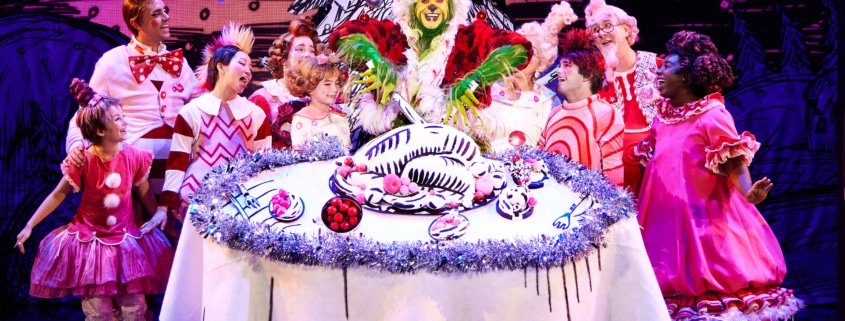 A green furry creature dressed in a santa suit carves into an illustrated turkey on top of a large, round table. Other colorful actors in pink costumes surround the table
