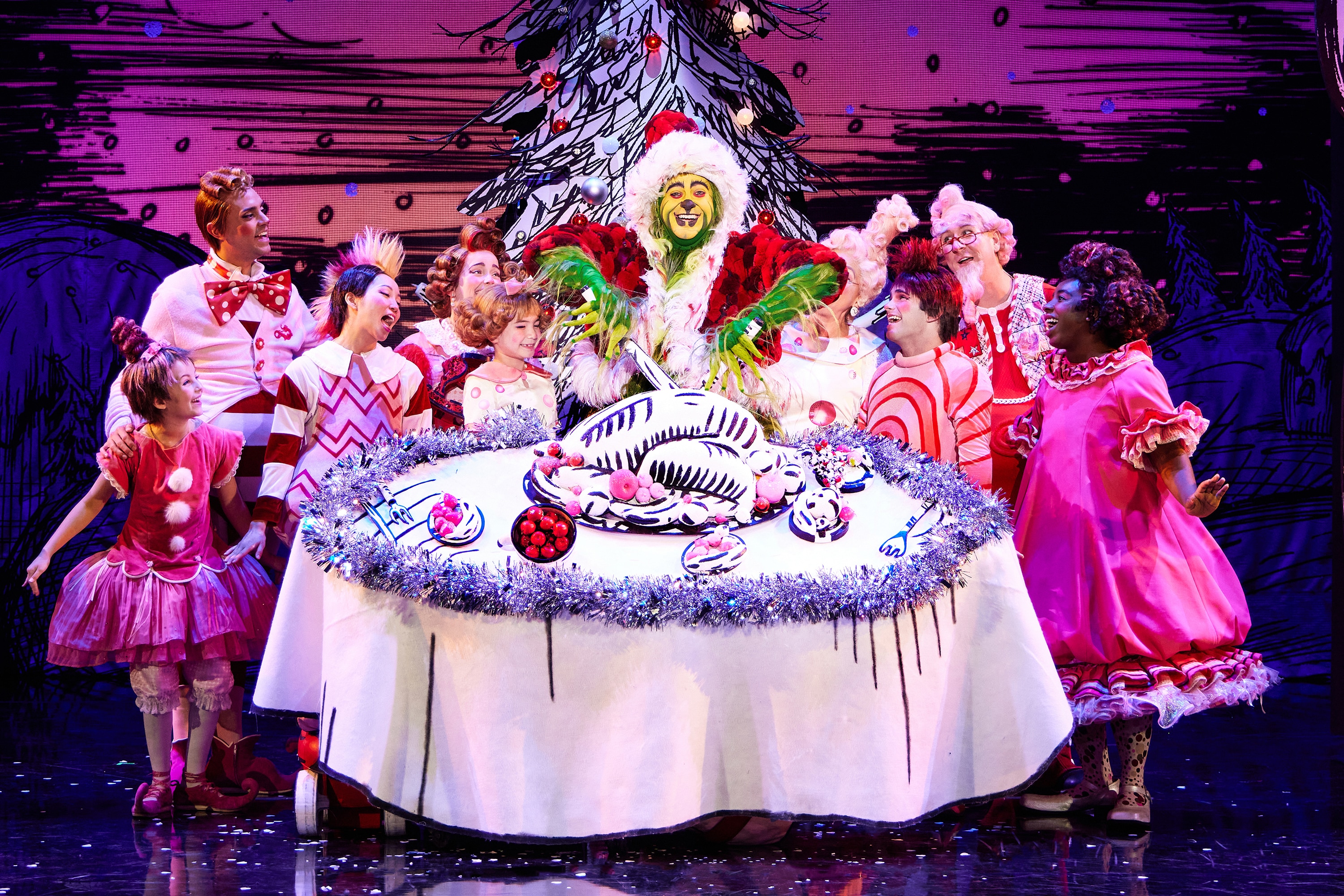 A green furry creature dressed in a santa suit carves into an illustrated turkey on top of a large, round table. Other colorful actors in pink costumes surround the table