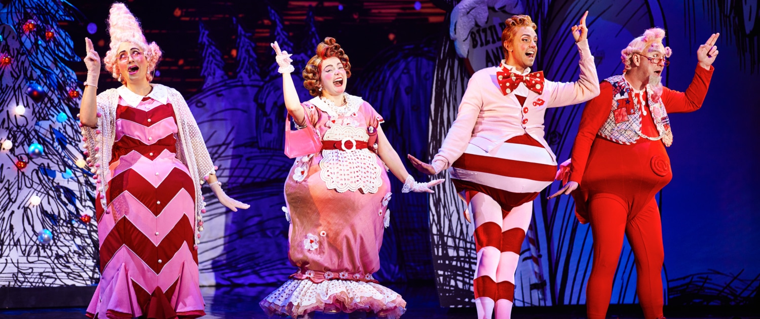 Four actors dressed in colorful, whimsical costumes performing on stage with a festive backdrop.
