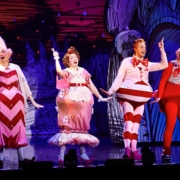 Four actors dressed in colorful, whimsical costumes performing on stage with a festive backdrop.