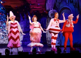 Four actors dressed in colorful, whimsical costumes performing on stage with a festive backdrop.