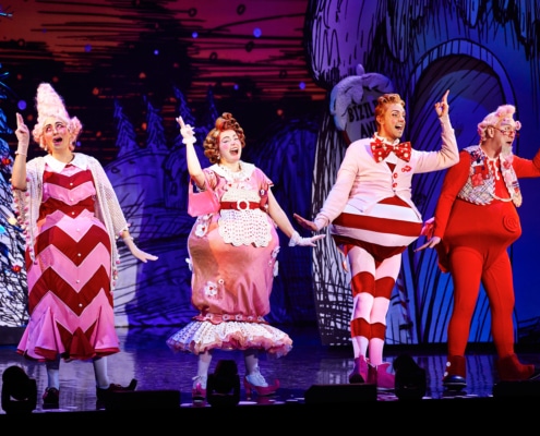 Four actors dressed in colorful, whimsical costumes performing on stage with a festive backdrop.