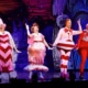 Four actors dressed in colorful, whimsical costumes performing on stage with a festive backdrop.