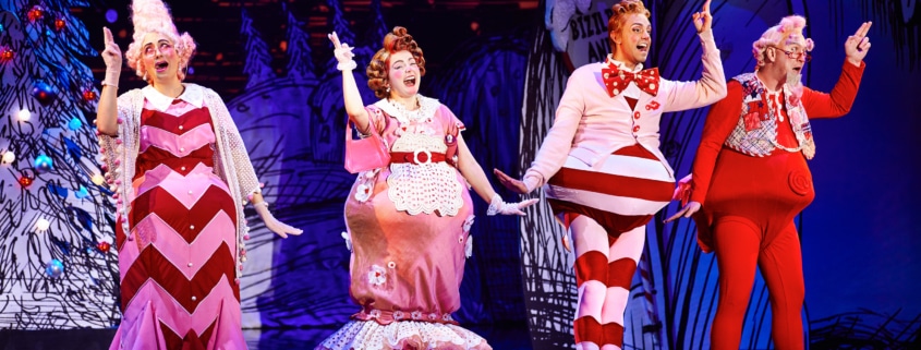 Four actors dressed in colorful, whimsical costumes performing on stage with a festive backdrop.