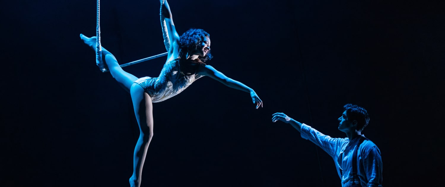 Ariel performers on a trapeze illuminated by blue light on a black background