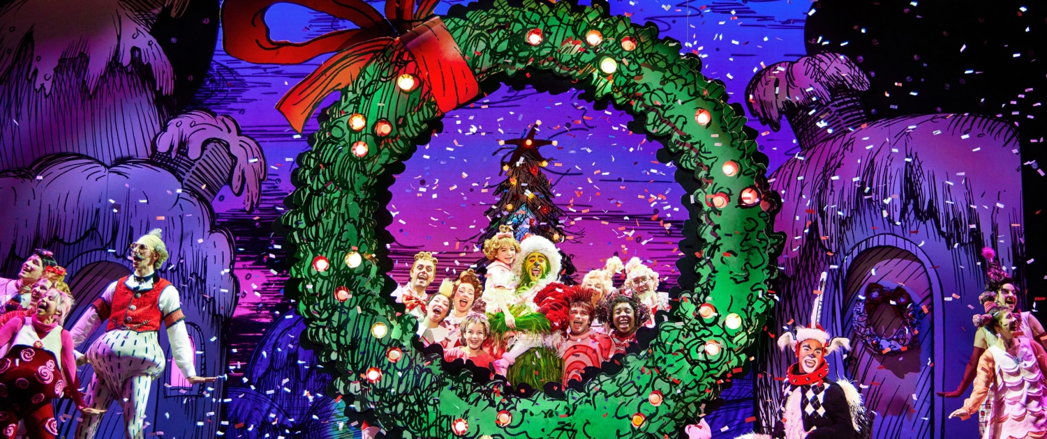 A festive stage performance with actors dressed in colorful costumes, centered around a large green wreath with a red bow, set against a vibrant backdrop of decorated trees and houses.