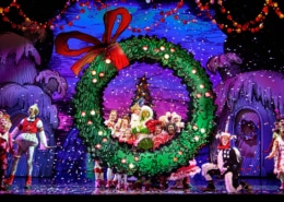 A festive stage performance with actors dressed in colorful costumes, centered around a large green wreath with a red bow, set against a vibrant backdrop of decorated trees and houses.