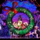 A festive stage performance with actors dressed in colorful costumes, centered around a large green wreath with a red bow, set against a vibrant backdrop of decorated trees and houses.