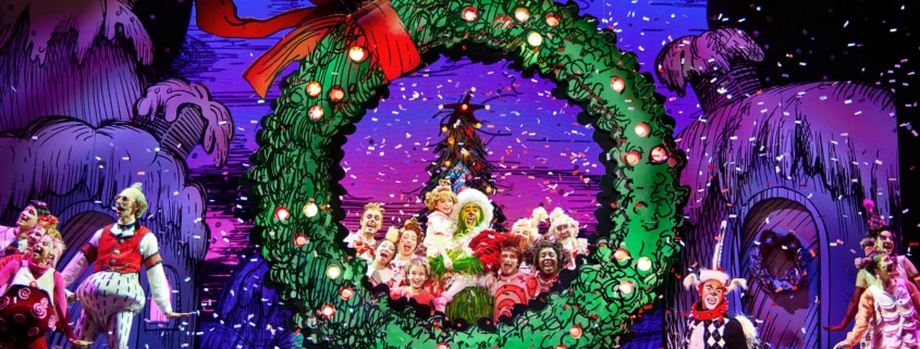 A festive stage performance with actors dressed in colorful costumes, centered around a large green wreath with a red bow, set against a vibrant backdrop of decorated trees and houses.