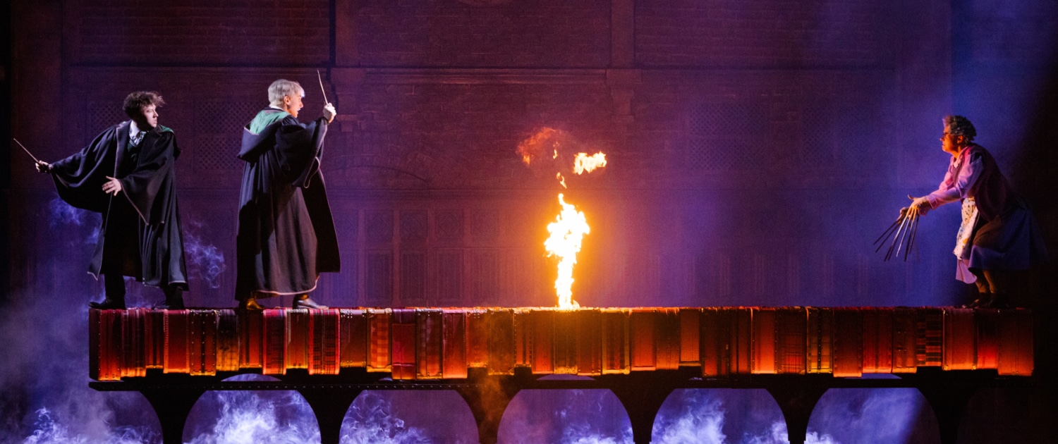 Two boys walk across a dimly lit bridge with a flame in the center while a witch blocks their path.