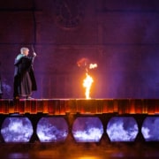 Two boys walk across a dimly lit bridge with a flame in the center while a witch blocks their path.