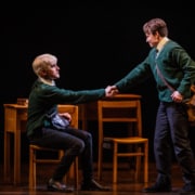 Two boys (one seated and one standing) shake hands in front of a table and chairs.