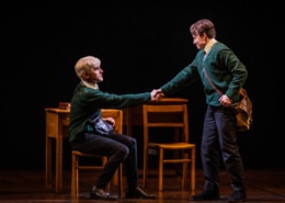 Two boys (one seated and one standing) shake hands in front of a table and chairs.