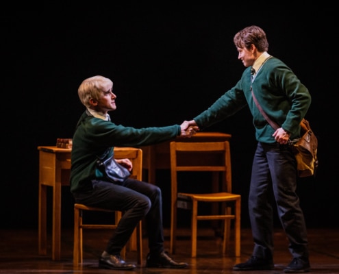 Two boys (one seated and one standing) shake hands in front of a table and chairs.