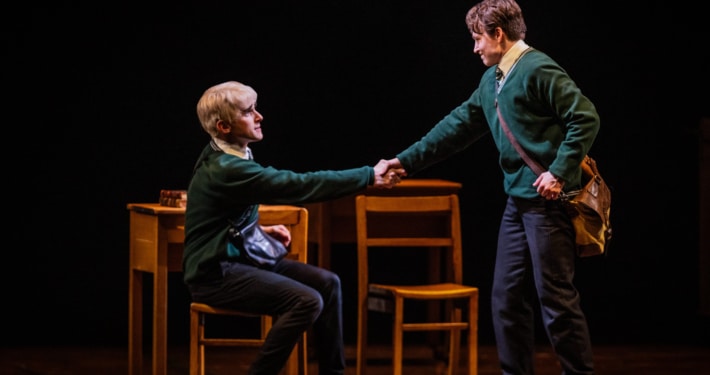 Two boys (one seated and one standing) shake hands in front of a table and chairs.