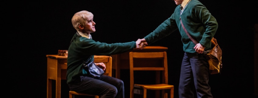 Two boys (one seated and one standing) shake hands in front of a table and chairs.