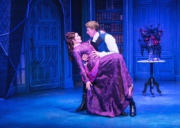 Stage scene with two actors in period costumes; one dips the other dramatically against a blue ornate backdrop.