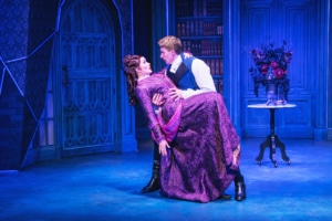 Stage scene with two actors in period costumes; one dips the other dramatically against a blue ornate backdrop.