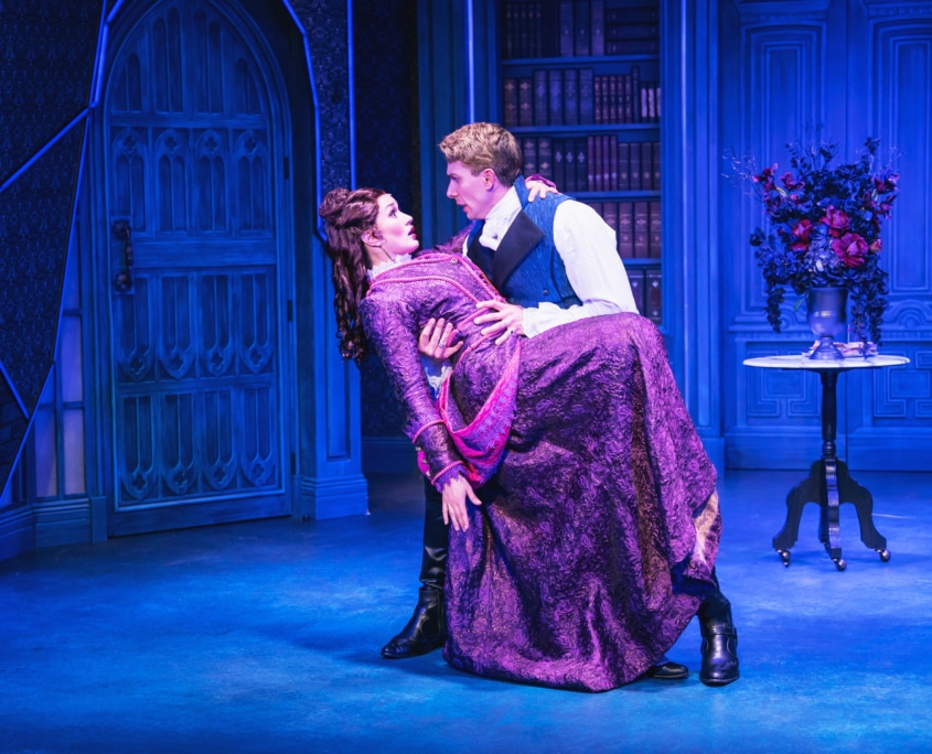 Stage scene with two actors in period costumes; one dips the other dramatically against a blue ornate backdrop.
