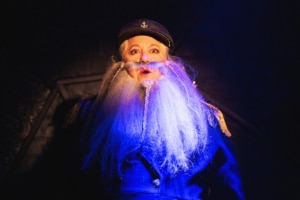 Person in a dark blue captain-style coat with gold shoulder accents and a long white beard, wearing a peaked cap under dramatic lighting.