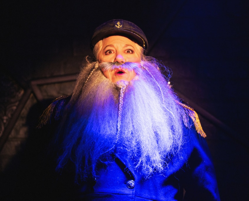 Person in a dark blue captain-style coat with gold shoulder accents and a long white beard, wearing a peaked cap under dramatic lighting.