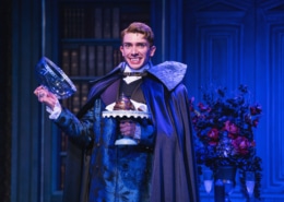 Actor in ornate blue costume and cape smiles, showing vampire fangs, and holds a glass dome with dessert on a blue-lit stage.