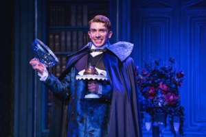 Actor in ornate blue costume and cape smiles, showing vampire fangs, and holds a glass dome with dessert on a blue-lit stage.