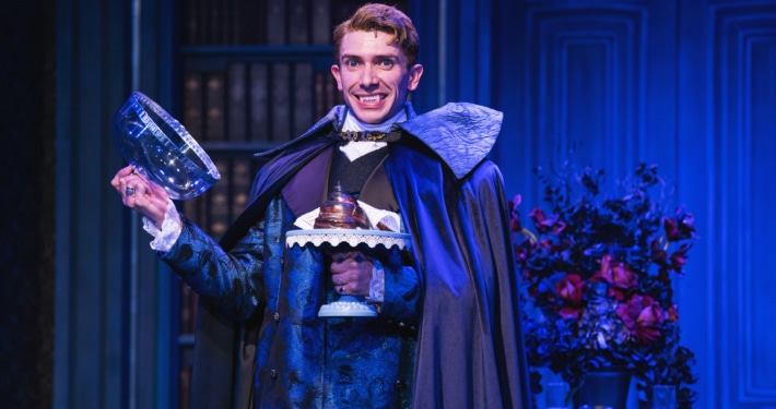 Actor in ornate blue costume and cape smiles, showing vampire fangs, and holds a glass dome with dessert on a blue-lit stage.