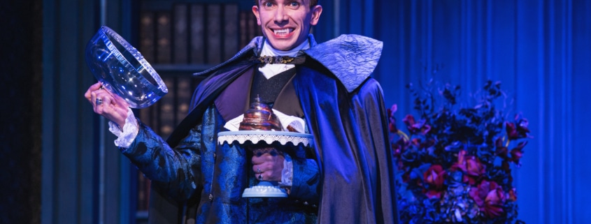 Actor in ornate blue costume and cape smiles, showing vampire fangs, and holds a glass dome with dessert on a blue-lit stage.