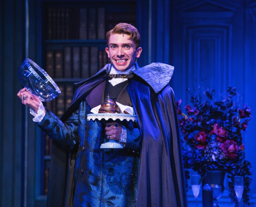 Actor in ornate blue costume and cape smiles, showing vampire fangs, and holds a glass dome with dessert on a blue-lit stage.