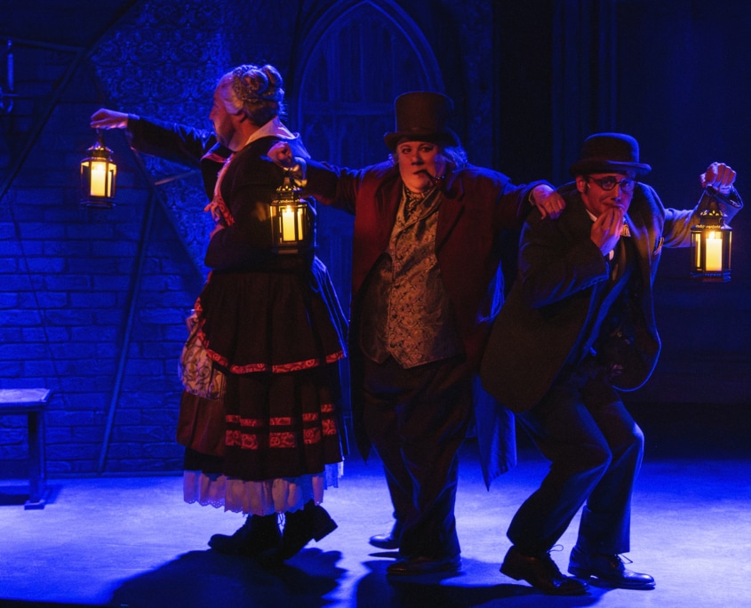 Three actors in 19th-century costumes hold lanterns on a dimly lit stage with gothic scenery.