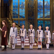 A photo of a group of seven children lined up by height at the bottom of a staircase, wearing outfits that resemble sailor attire. An adult couple stands to their left.