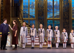 A photo of a group of seven children lined up by height at the bottom of a staircase, wearing outfits that resemble sailor attire. An adult couple stands to their left.