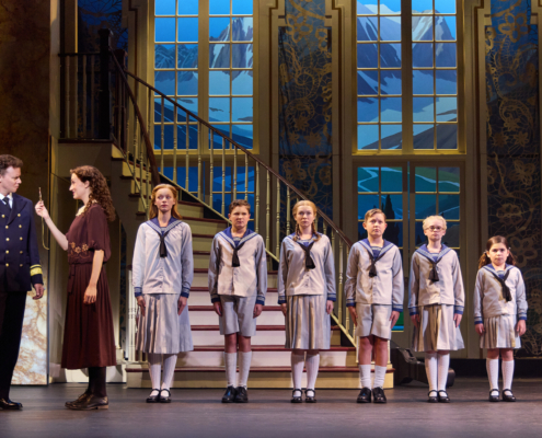 A photo of a group of seven children lined up by height at the bottom of a staircase, wearing outfits that resemble sailor attire. An adult couple stands to their left.