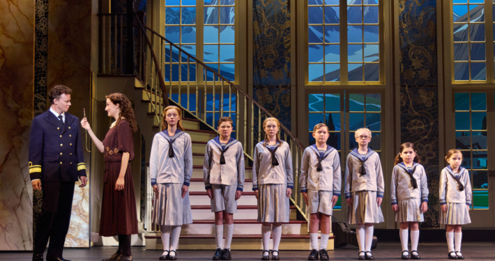 A photo of a group of seven children lined up by height at the bottom of a staircase, wearing outfits that resemble sailor attire. An adult couple stands to their left.
