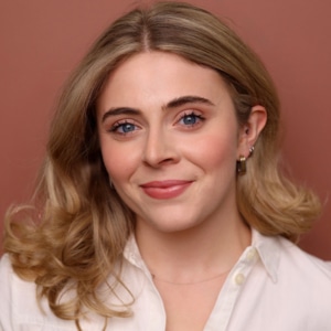 Headshot of an actress on a rust-colored background wearing a white blouse with long blond hair and a grin.