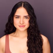 Headshot of an actress on a dark gray background wearing a coral tank top with long black wavy hair.