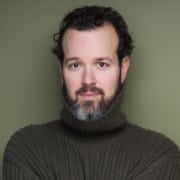 Headshot of actor with short, curly dark brown hair, beard, and dark turtleneck in front of green background