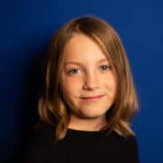 Headshot of young actress with shoulder-length brown hair and black crew-neck shirt in front of blue background.