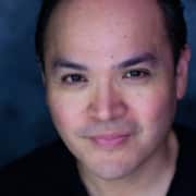 Headshot of an actor on a dark gray background wearing a black t-shirt with dark eyebrows.