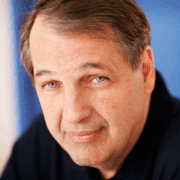 Headshot of actor with short brown hair, blue eyes, and black shirt in front of blue background