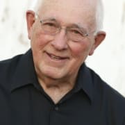 Headshot of actor with white hair, glasses, and black shirt.