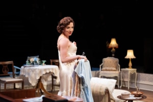 A woman in a satin nightgown stands in a vintage-style room holding a bundle of clothes. The room features antique furniture, lace-covered tables, and warm lighting from table lamps, evoking a domestic, mid-century Southern setting.