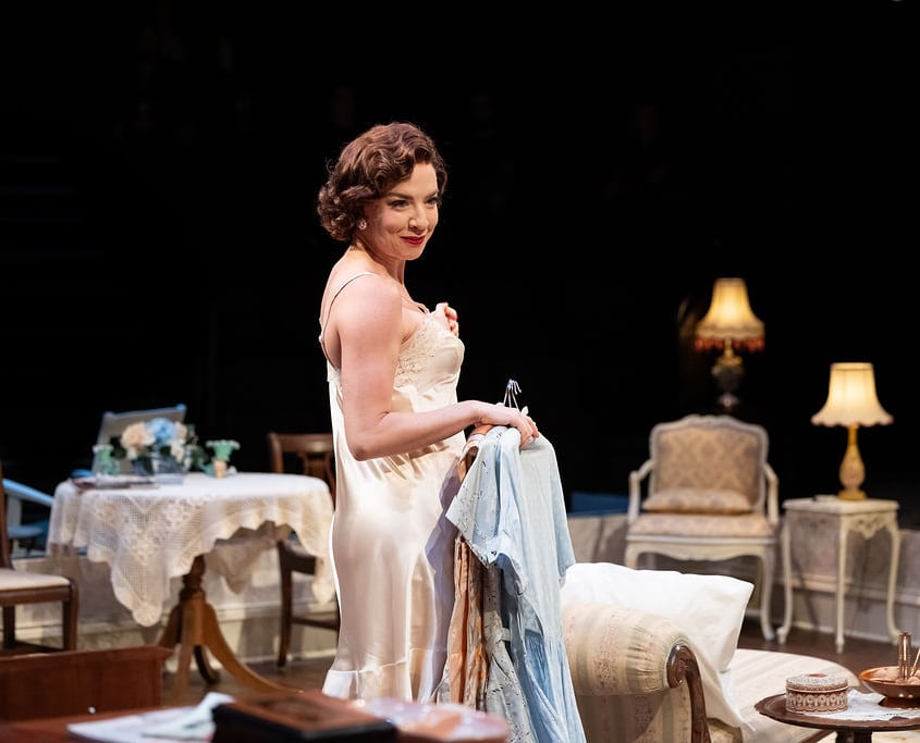 A woman in a satin nightgown stands in a vintage-style room holding a bundle of clothes. The room features antique furniture, lace-covered tables, and warm lighting from table lamps, evoking a domestic, mid-century Southern setting.