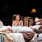 A man lies on a bed with his bandaged foot elevated, while a woman sits beside him, appearing to speak intently. The room is filled with period furniture and soft lighting, emphasizing an intimate moment.