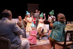 Children wearing party hats perform or dance in front of seated adults. A birthday cake with lit candles sits on a table, suggesting a celebratory scene within a family gathering.