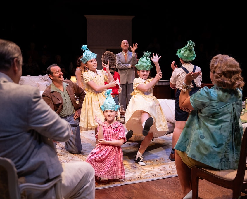 Children wearing party hats perform or dance in front of seated adults. A birthday cake with lit candles sits on a table, suggesting a celebratory scene within a family gathering.